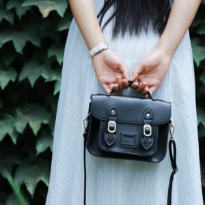A woman in a striped dress holds a black leather handbag with lush green leaves in the background.